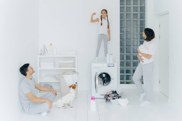 強い陽気な女の子は腕を上げ、足を見せ、両親を楽しませ、洗濯服の後に休憩し、ペット、洗濯機、洗浄剤で広々とした白い洗濯室でポーズをとっています。家事・家事 - 写真・画像