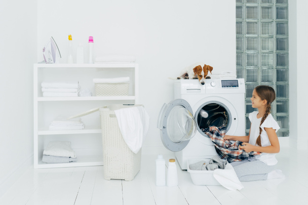 Housekeeping, children and domestic chores concept. Happy kid unloads washing machine, puts clean washed clothes in basin, curious dog looks from above, lies on washer in laundry room at home - Photo, Image