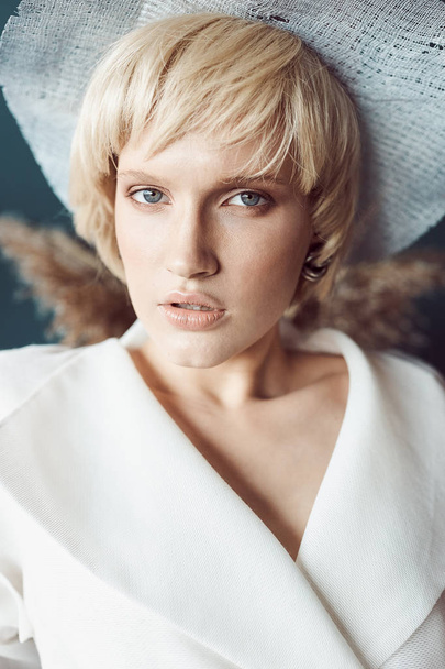 portrait of beautiful girl with blue eyes and short blonde hair, she has professional nude makeup, white hat on head and big earrings in ears, she looks in the camera   - 写真・画像