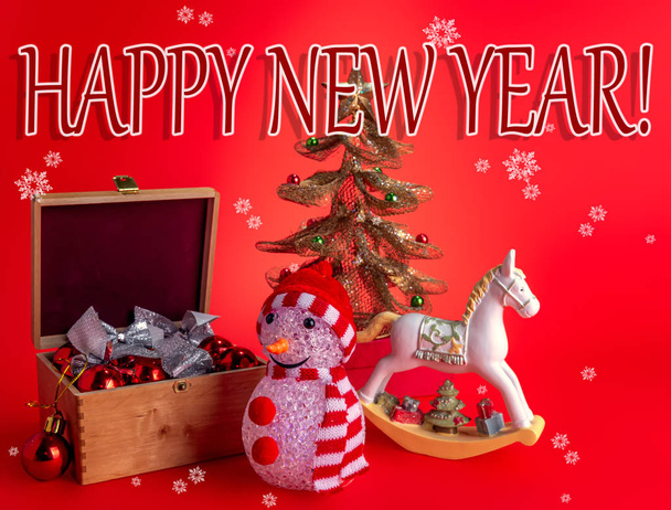 Feestelijke compositie met aangename wensen, een sneeuwpop, kerstspeelgoed en een paard op een rode achtergrond met sneeuwvlokken. - Foto, afbeelding