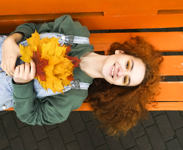 Beautiful young redhead woman lying on bench in sunny autumn day. top view. cute happy beautiful girl lying outdoors. Looking at camera. smiley face.ginger female hold in hands bouquet of maple leaves .  - Photo, Image