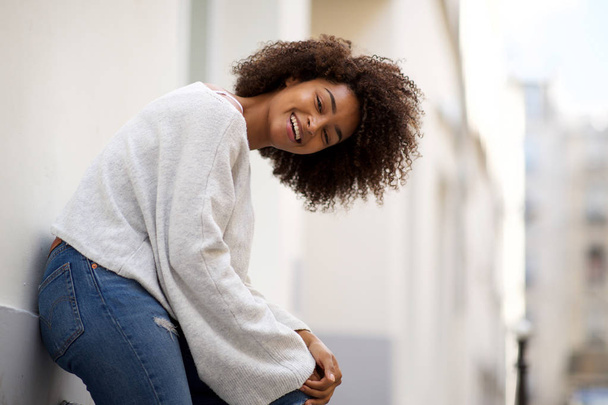 Portrait latéral de heureuse jeune femme afro-américaine appuyée contre le mur à l'extérieur
 - Photo, image