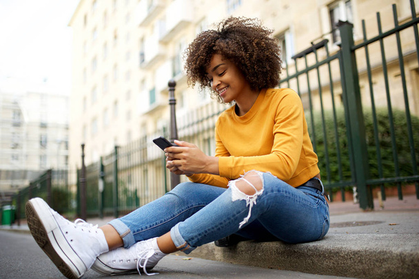 Πορτρέτο της δροσερό νεαρή Αφροαμερικανή γυναίκα κάθεται έξω στο δρόμο με το κινητό τηλέφωνο - Φωτογραφία, εικόνα