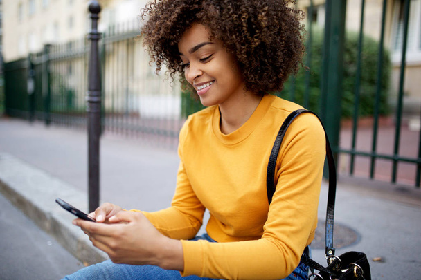 Портрет улыбающейся африканской американки, смотрящей на мобильный телефон в городе
 - Фото, изображение