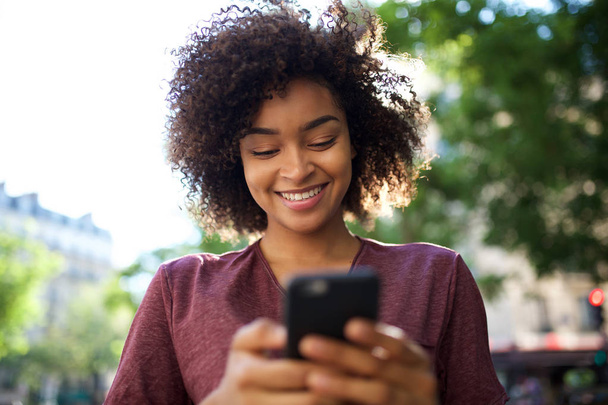 Κοντινό πλάνο πορτρέτο της χαμογελαστής νεαρής Αφροαμερικανής γυναίκας που κοιτάζει κινητό τηλέφωνο σε εξωτερικούς χώρους - Φωτογραφία, εικόνα