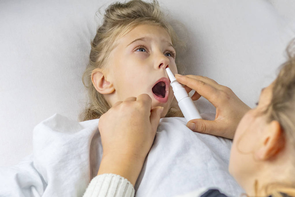 Ágy. Egy fehér hajú kislány nyitott szájjal fekszik. anya beadja az injekciót az orrába. - Fotó, kép
