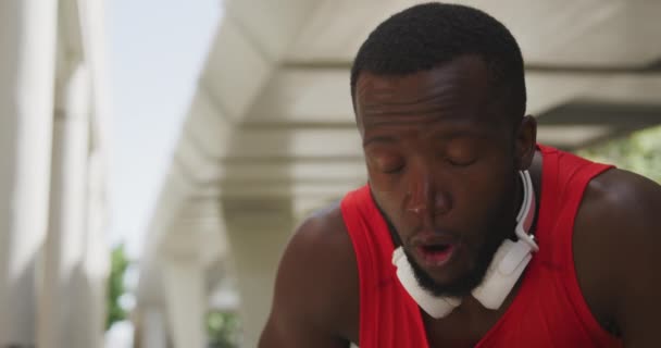 Front view close up of an athletic African American man in sportswear with headphones around his neck taking a break, catching his breath and mopping his brow, under a bridge while training on a sunny day in the city, slow motion - Footage, Video