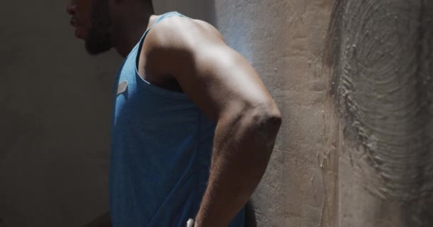 Side view close up of an athletic African American man in sportswear taking a break, leaning against a wall and drinking water under a bridge while training on a sunny day in the city, slow motion - Footage, Video