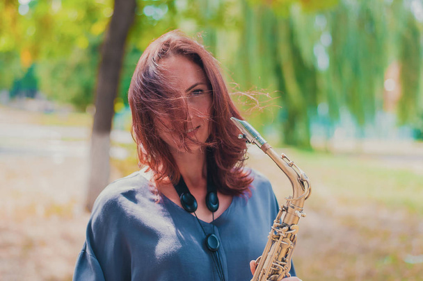 kaunis punatukkainen tyttö sinisessä takissa saksofonin kanssa tuulisella säällä
 - Valokuva, kuva