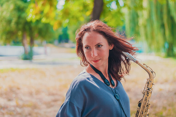 Близкий портрет молодой рыжеволосой женщины в зеленом парке с саксофоном
 - Фото, изображение