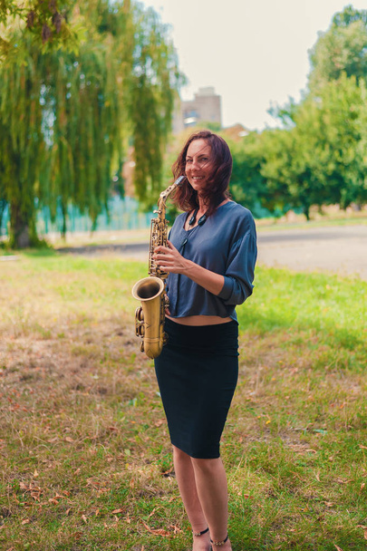 słodkie młody ruda kobieta w niebieski bluzka i czarny spódnica pozowanie z saksofon w park - Zdjęcie, obraz