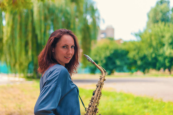 zrzavá mladá dívka v modré bundě se saxofonem usmívající se v parku - Fotografie, Obrázek