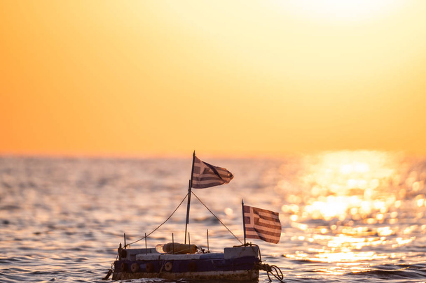 Yaz denizinde Yunan bayrağı taşıyan oyuncak tekne, arka planda güzel bir gün doğumu, seyahat konsepti  - Fotoğraf, Görsel