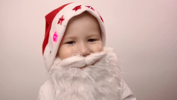 three-year-old caucasian kid with artificial beard in Santa Claus hat with blinking red lights is smiling and laughing on white background. Funny child costume for celebrating new year and christmas - Filmmaterial, Video