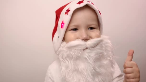 three-year-old caucasian kid with artificial beard in Santa Claus hat with blinking red lights smiles, laughs, pulls approving thumbs up sign on white background. Funny new year or christmas costume - Filmagem, Vídeo