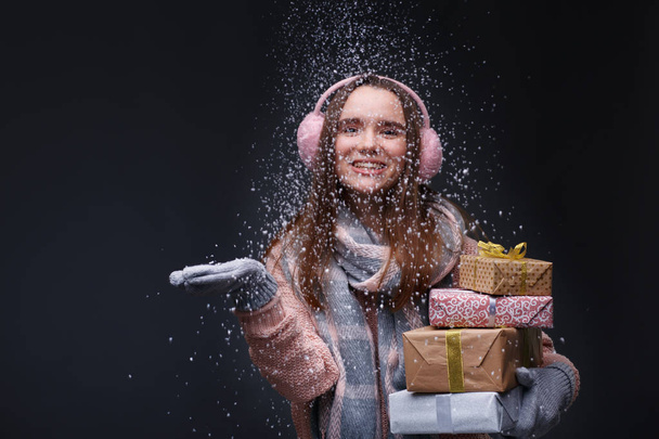 Jong mooi lachend meisje in gebreide trui en roze pluizige oorkappen met cadeaudoosjes en blazende witte sneeuwvlokken op een grijze achtergrond. Wintervakantie, Kerstmis, Nieuwjaarsconcept.  - Foto, afbeelding