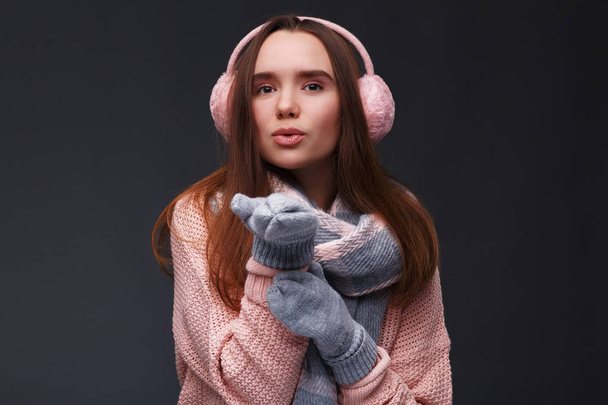 Portrait of young beautiful smiling girl in knitting pullover, mittens and pink fluffy earmuffs on a dark background. Xmas model with long straight hair. Winter holidays, Christmas, New Year concept. - Photo, Image