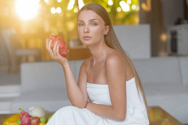 νεαρή γυναίκα τυλιγμένη σε λευκή πετσέτα κάθεται με εξωτικά φρούτα στο σπα - Φωτογραφία, εικόνα