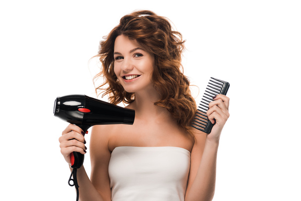 feliz chica rizada sosteniendo secador de pelo y cepillo aislado en blanco
  - Foto, Imagen