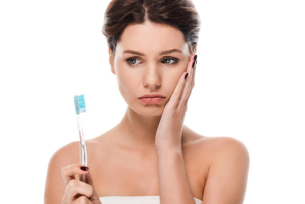 traurige junge Frau berührt Gesicht, während sie Zahnbürste isoliert auf weiß hält  - Foto, Bild