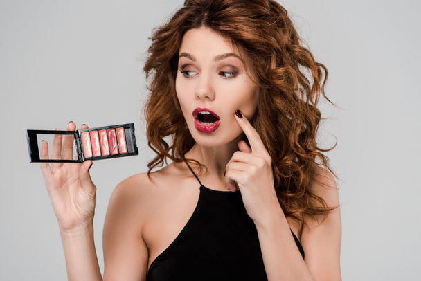 shocked curly woman looking at set of lip gloss isolated on grey  - Fotó, kép