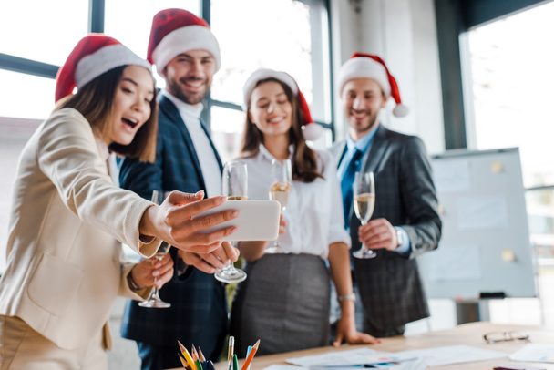 Mutlu, çok kültürlü iş kadınlarının ve Noel Baba şapkalı iş adamlarının ofiste şampanya bardaklarıyla selfie çektikleri seçici odak noktası.   - Fotoğraf, Görsel