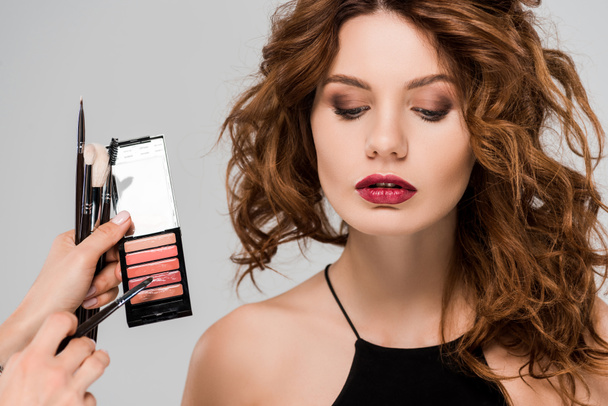 cropped view of makeup artist holding lip gloss near beautiful woman isolated on grey  - 写真・画像