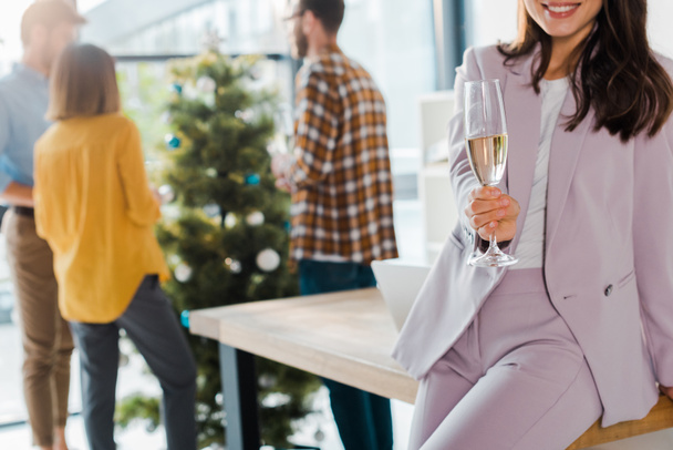 cropped view of businesswoman holding champagne glass near coworkers and christmas tree  - 写真・画像