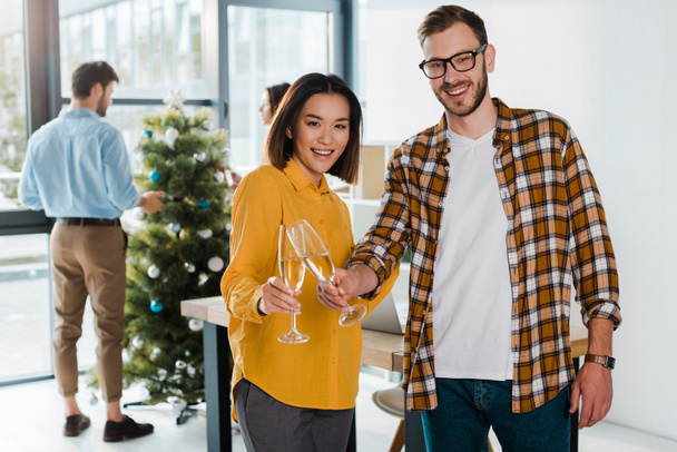 valikoiva painopiste iloinen liikemies ja aasialainen liikenainen paahtaminen samppanjalasit lähellä kollegoita ja joulukuusi
  - Valokuva, kuva