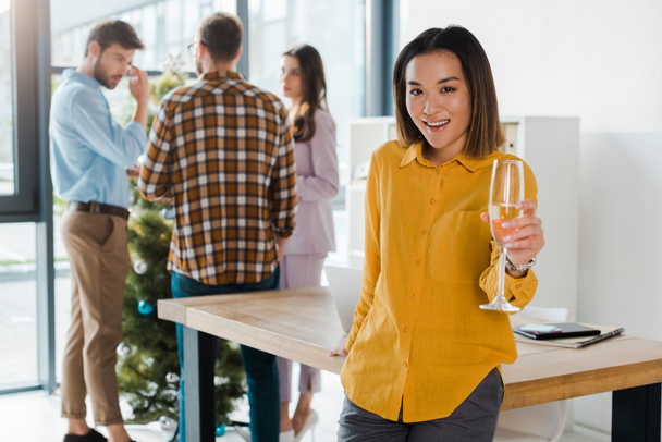 selective focus of happy asian woman holding champagne glass near coworkers and christmas tree in office  - Photo, Image