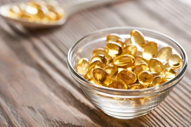 selective focus of golden fish oil capsules in glass bowl and spoon on wooden table - Fotoğraf, Görsel