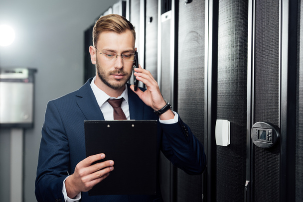 handsome businessman talking on smartphone while looking at clipboard in server room  - Photo, Image
