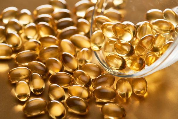 close up view of shiny fish oil capsules scattered from glass bowl on golden background - Photo, Image