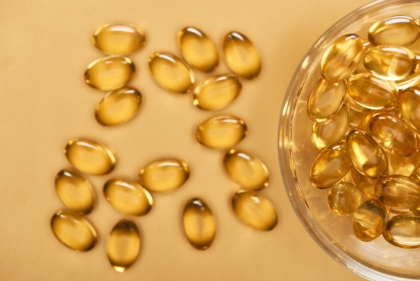 top view of shiny golden fish oil capsules scattered from glass bowl on yellow background - Photo, Image