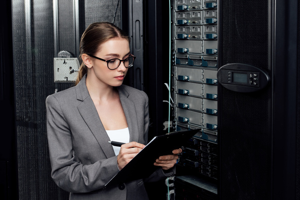 businesswoman in glasses holding clipboard while writing in data center  - Foto, imagen