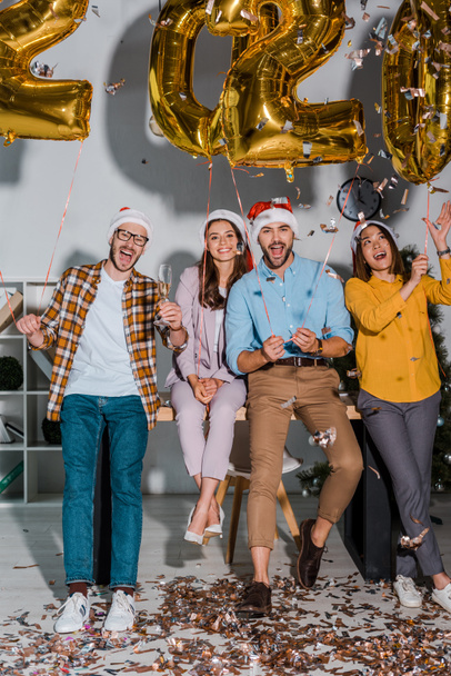happy businessmen and multicultural businesswomen in santa hats with champagne glasses near falling confetti and balloons with numbers  - Zdjęcie, obraz