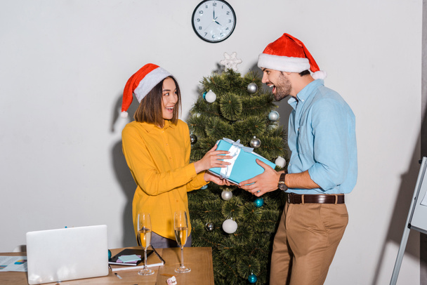 glücklicher bärtiger Geschäftsmann überreicht Geschenk an asiatische Geschäftsfrau mit Weihnachtsmann-Hut  - Foto, Bild