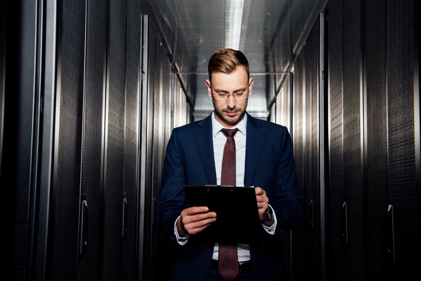 handsome businessman in glasses looking at clipboard near server racks  - Photo, Image