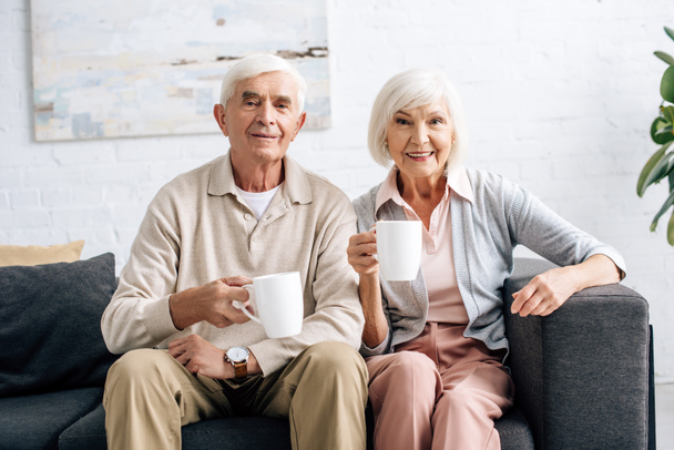 smiling husband and wife holding cups and sitting on sofa in apartment  - Foto, immagini