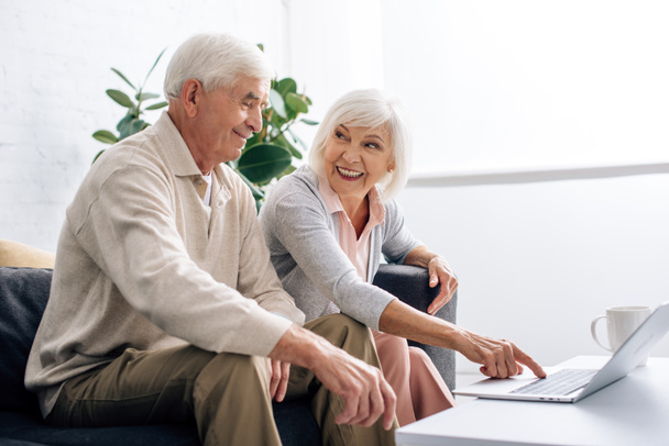 man en lachende vrouw met behulp van laptop en zitten op de bank in appartement  - Foto, afbeelding