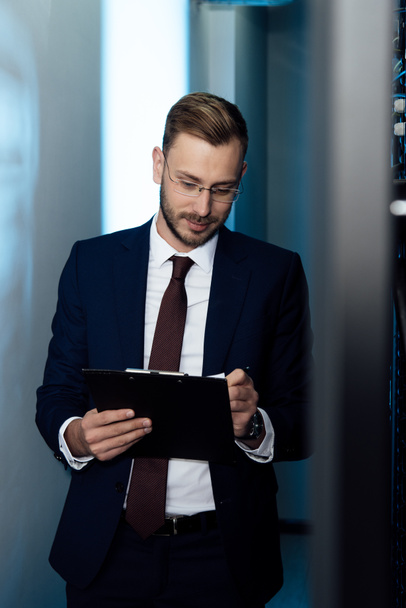 bearded businessman in glasses holding pen and clipboard in data center  - Foto, afbeelding