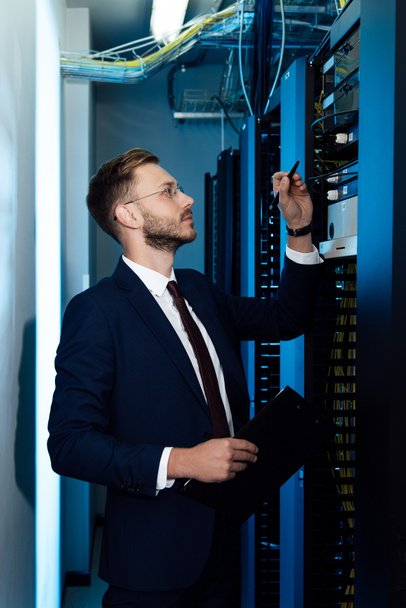 profile of handsome businessman in glasses holding pen and clipboard while looking at server rack  - Foto, immagini