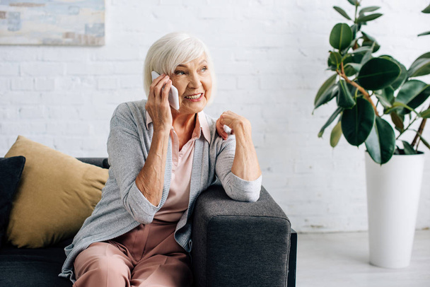 smiling senior woman sitting on sofa and talking on smartphone in apartment  - Foto, imagen