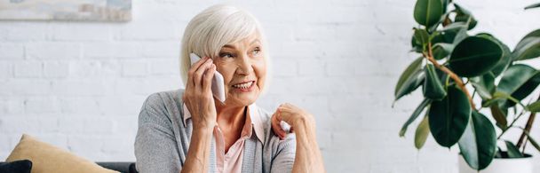 panoramic shot of smiling senior woman talking on smartphone in apartment  - Foto, Imagen