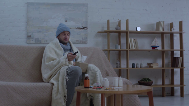 nemocný muž sedí u stolu s léky, kýchání a sledování televize  - Záběry, video
