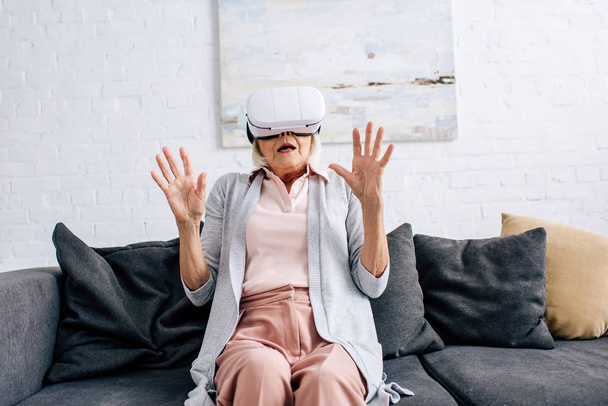 потрясенная пожилая женщина с гарнитурой виртуальной реальности, сидящая на диване в квартире
  - Фото, изображение