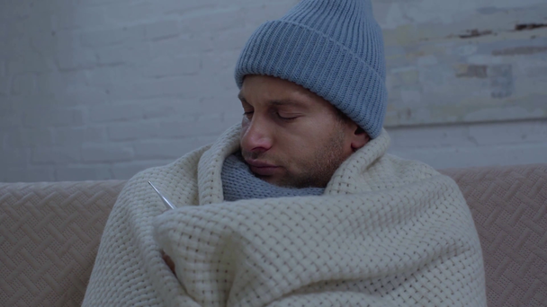 Kranker Mann mit warmer Mütze in Decke gewickelt und Temperatur gemessen - Filmmaterial, Video