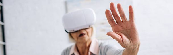 tiro panorâmico de mulher idosa em fone de ouvido realidade virtual com mão estendida no apartamento
  - Foto, Imagem