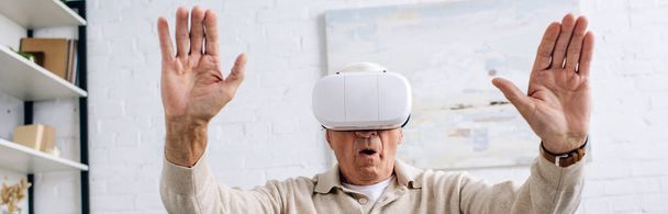 панорамный снимок пожилого человека в гарнитуре виртуальной реальности с протянутыми руками в квартире
  - Фото, изображение