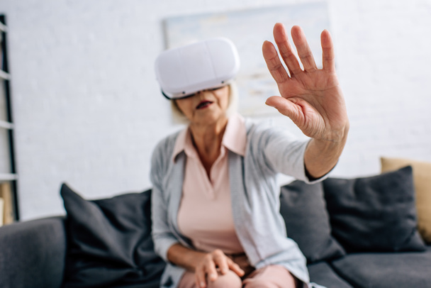 selektywna ostrość seniorki w wirtualnej rzeczywistości zestaw słuchawkowy z wyciągniętą ręką w mieszkaniu  - Zdjęcie, obraz
